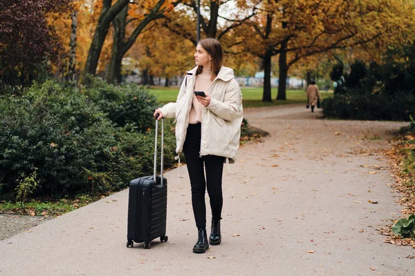 Pretty casual girl in down jacket thoughtfully walking through city park with suitcase — Stock Photo, Image