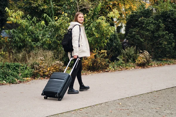 Gadis cantik tersenyum di bawah jaket senang berjalan melalui taman kota dengan koper dan ransel — Stok Foto