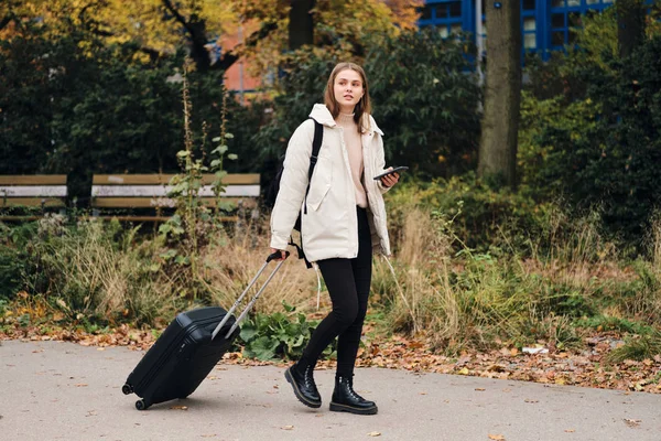Привлекательная случайная девушка в пуховике мечтательно прогуливается по городскому парку с чемоданом и рюкзаком — стоковое фото