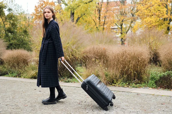 Красивая стильная девушка в пальто тянет свой багаж сумка задумчиво прогуливаясь по городскому парку — стоковое фото