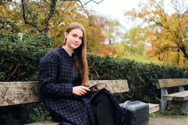 Привлекательная случайная девушка с планшетным сидением с чемоданом на скамейке в городском парке — стоковое фото