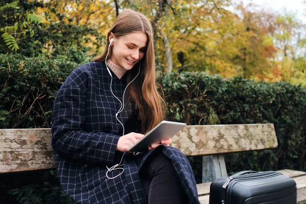 Pěkně ležérní dívka ve sluchátkách šťastně pomocí tabletu sedí s kufrem na lavičce v městském parku — Stock fotografie
