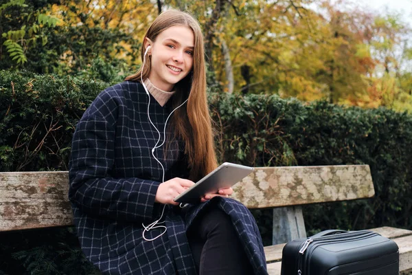 Pěkně usměvavá dívka s kufrem šťastně pomocí tabletu odpočívá na lavičce v městském parku — Stock fotografie