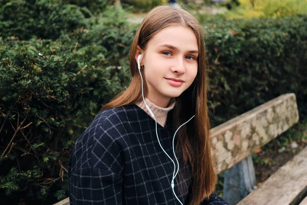 Retrato de bela menina sorridente em fones de ouvido olhando alegremente na câmera no banco no parque — Fotografia de Stock