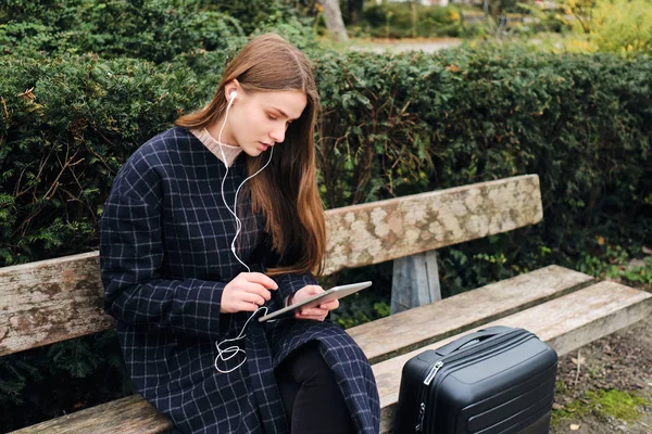 街の公園のベンチにスーツケースと一緒に座ってタブレットを使用して思慮深くイヤホンの美しいカジュアルな女の子 — ストック写真