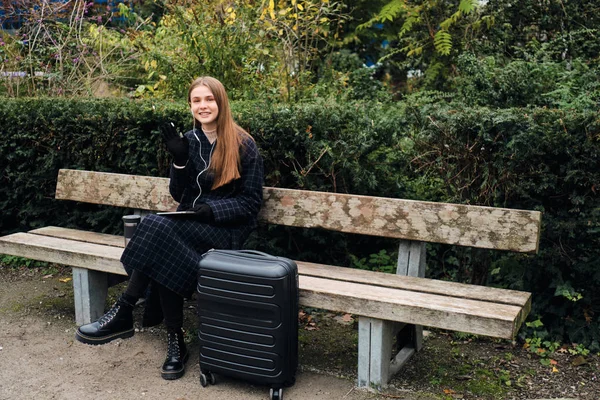 Fröhliches stilvolles Mädchen winkt fröhlich mit Hallo-Geste in die Kamera, sitzend mit Koffer auf Bank im Park — Stockfoto