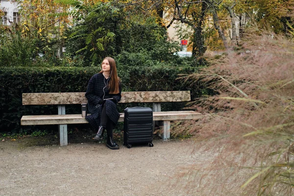 Schöne stilvolle Mädchen mit Koffer verträumt wartet auf Bank im Stadtpark — Stockfoto