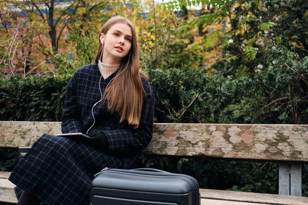 Attraktives stylisches Mädchen, das verträumt mit Gepäck auf Bank im Stadtpark wartet — Stockfoto