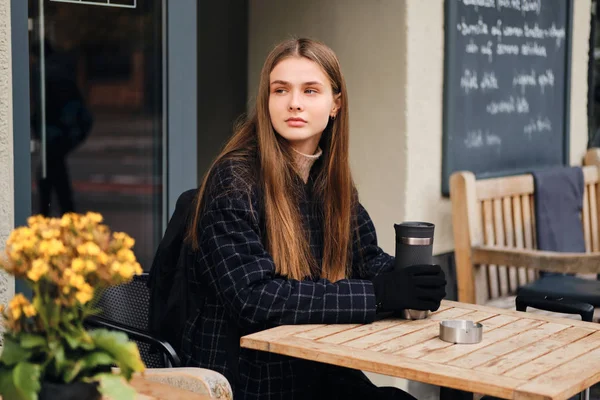 Menina elegante bonita segurando copo termo pensativo olhando para longe no acolhedor café de rua — Fotografia de Stock