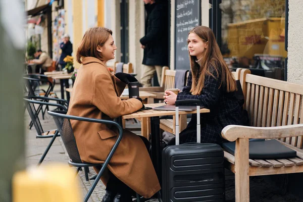2人の美しいスタイリッシュな女の子が夢のようにスーツケースと屋外カフェで一緒に休んで話をする — ストック写真