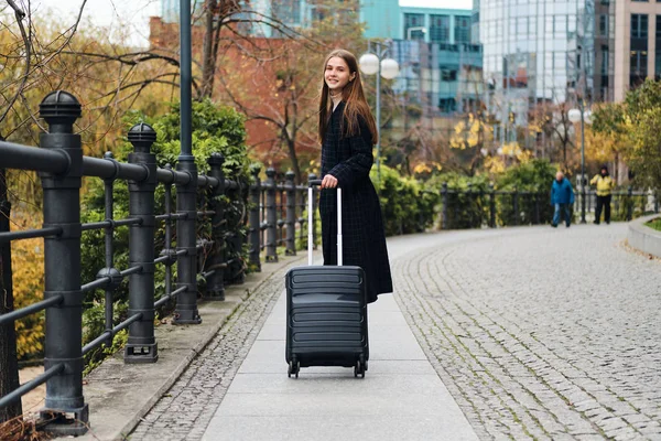 Belle fille élégante en manteau avec sac à bagages regardant joyeusement dans la caméra sur la rue de la ville — Photo