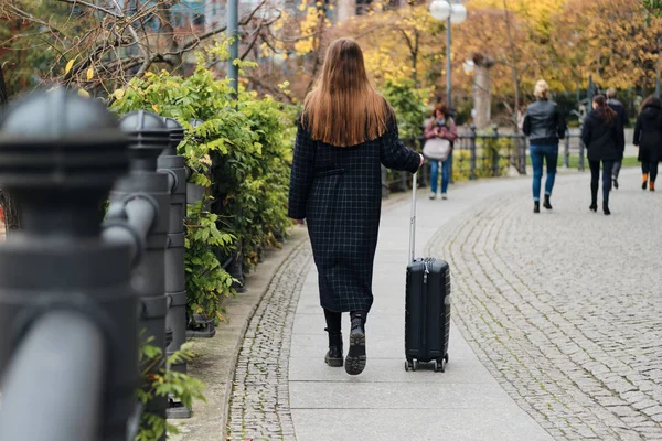 Widok z tyłu casual girl w płaszczu spacery po ulicy z walizką — Zdjęcie stockowe