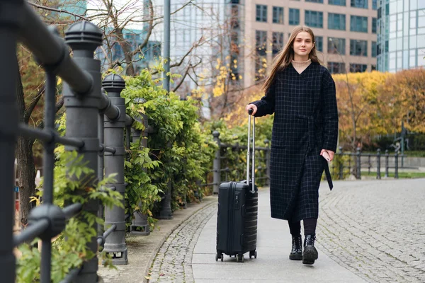 Hübsches stylisches Mädchen im Mantel, das verträumt mit einem Koffer durch die Stadt läuft — Stockfoto