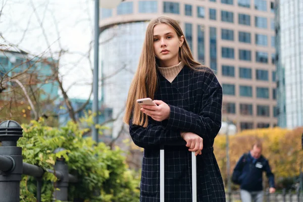Krásná ležérní dívka v kabátu pomocí mobilního telefonu stojící na ulici města s kufrem — Stock fotografie