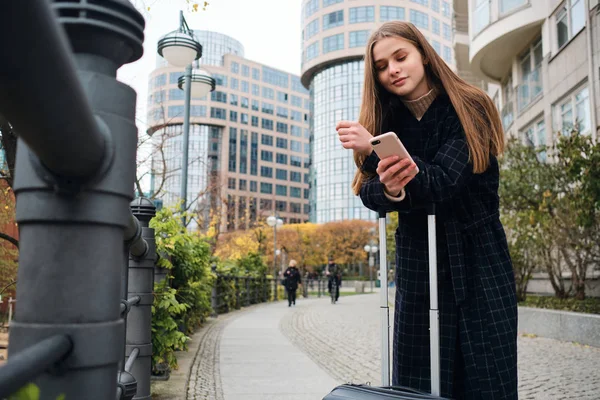 Menina sorrindo atraente no casaco alegremente usando celular de pé na rua da cidade com mala — Fotografia de Stock