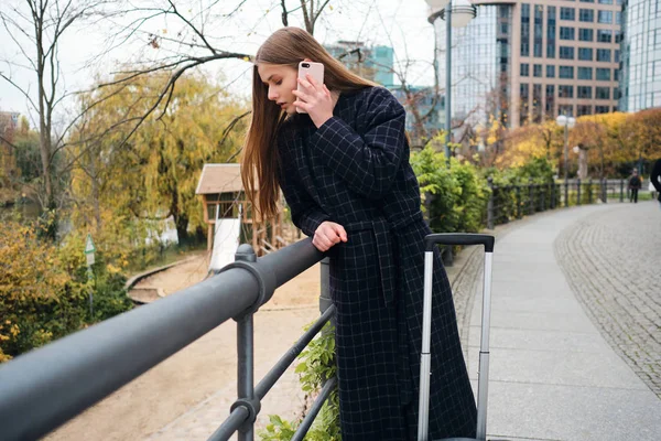 Красивая улыбающаяся девушка в пальто счастливо разговаривает по мобильному телефону в городском парке — стоковое фото