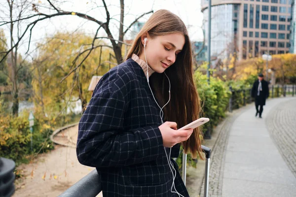 Menina casual atraente em fones de ouvido ouvir música alegremente no celular durante a caminhada no parque da cidade — Fotografia de Stock
