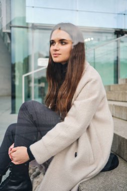 Çekici esmer kız, paltolu. Üzüntüyle başka yere bakıyor. Dışarıda merdivenlerde oturuyor.