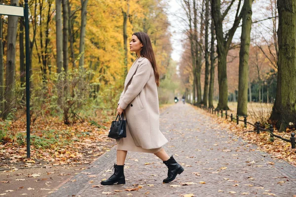 Vista lateral de chica morena atractiva en abrigo caminando por el hermoso parque de otoño solo — Foto de Stock