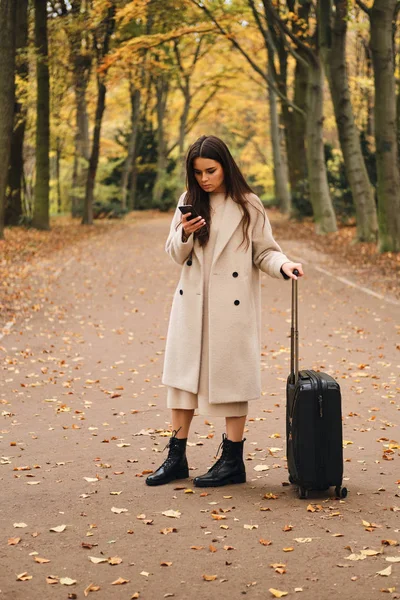 Muchacha atractiva con estilo en el abrigo intensamente utilizando el teléfono celular de pie en el hermoso parque de otoño con maleta — Foto de Stock