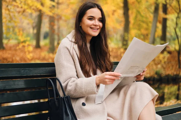 Красивая улыбающаяся брюнетка счастливо читает газету на скамейке запасных в осеннем городском парке — стоковое фото