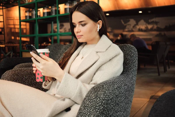 Красивая стильная деловая женщина в беспроводных наушниках уверенно использует сотовый телефон отдыха в современном кафе — стоковое фото