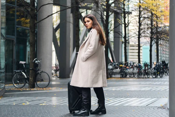 Atractiva chica con estilo en abrigo con confianza mirando a un lado de pie en la calle de la ciudad con poca maleta — Foto de Stock