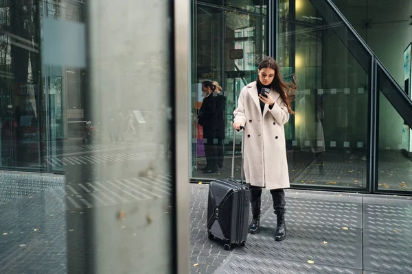 Jeune belle femme d'affaires en manteau à l'aide d'un téléphone portable attendant dans la rue avec valise — Photo