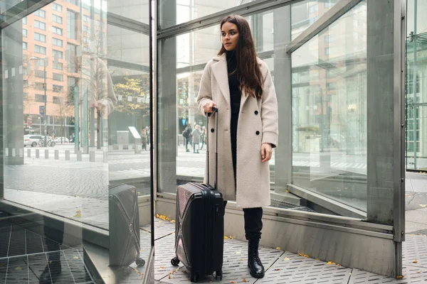 Junge stilvolle Geschäftsfrau im Mantel steht nachdenklich mit Koffer draußen — Stockfoto