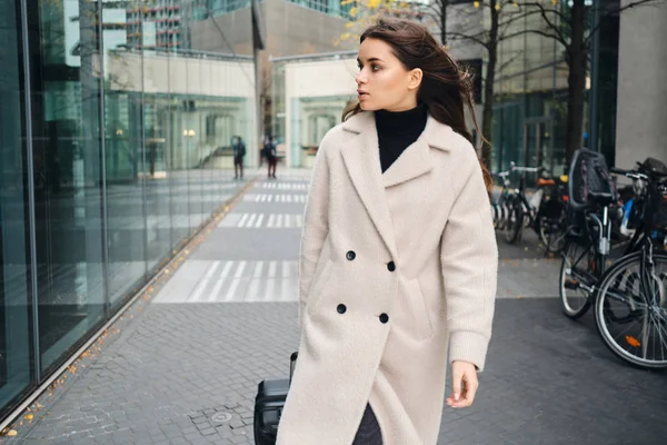 Joven mujer de negocios atractiva en abrigo caminando con confianza por la calle de la ciudad con la maleta — Foto de Stock