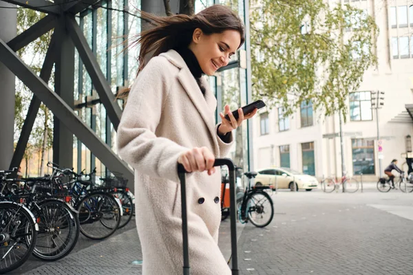 Junge hübsche Geschäftsfrau im Mantel nutzt fröhlich Handy, wenn sie mit Koffer durch die Straße läuft — Stockfoto