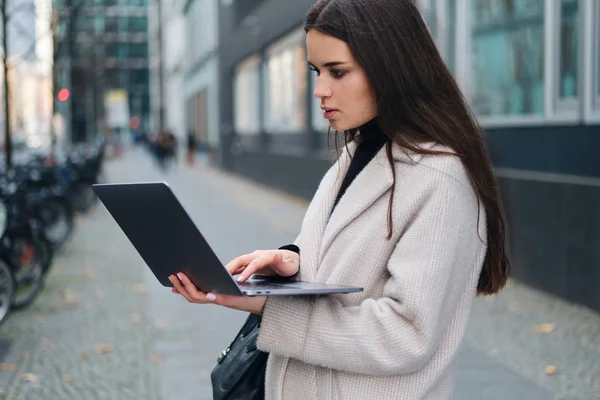 Vista lateral da jovem mulher de negócios elegante que trabalha intensamente no laptop na rua da cidade — Fotografia de Stock