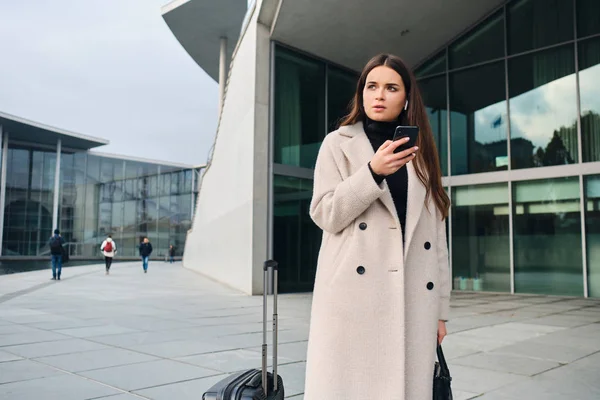 Wanita pengusaha bergaya muda bermantel berpikir menggunakan ponsel yang menunggu di jalan kota dengan koper — Stok Foto