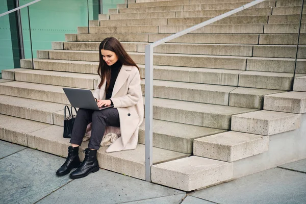 Молодая стильная деловая женщина счастливо работает на ноутбуке, сидя на лестнице — стоковое фото