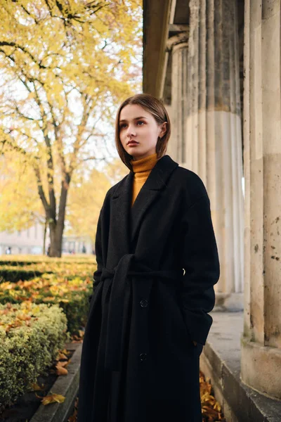 Menina elegante bonita em casaco preto atentamente olhando para longe na bela rua da cidade — Fotografia de Stock