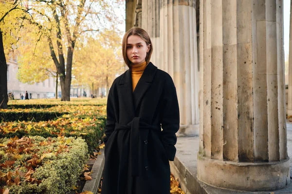 Ernstes stylisches Mädchen in schwarzem Mantel, das gespannt in die Kamera auf der schönen herbstlichen Stadtstraße blickt — Stockfoto
