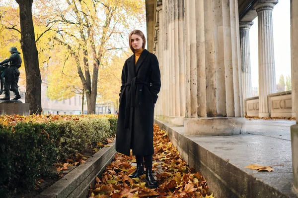 Attraktives, stylisches Mädchen in schwarzem Mantel blickt selbstbewusst in die Kamera auf der Stadtstraße voller gelber Blätter — Stockfoto