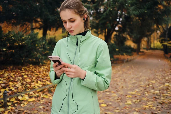 Attraente ragazza sportiva in auricolare utilizzando il cellulare dopo la corsa in accogliente parco autunnale — Foto Stock