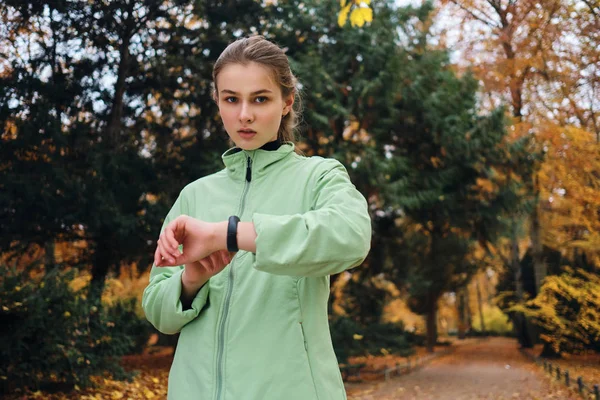 Bela menina esportiva verificando o progresso no relógio inteligente após correr no parque de outono — Fotografia de Stock