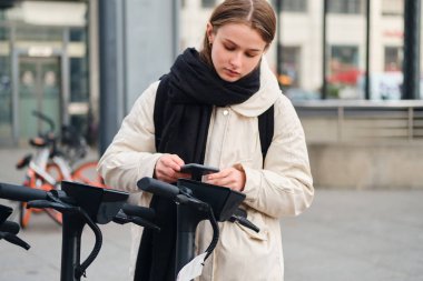 Çekici kız kendinden emin bir şekilde sokakta elektrikli scooter kiralamak için cep telefonu kullanıyor.