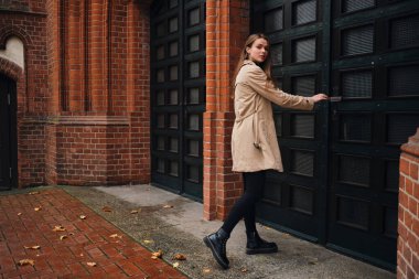 Trençkotlu güzel bir kız kameraya bakıyor caddede eski kapıyı açıyor.