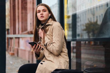 Kulaklıklı çekici bir kız cep telefonuyla dışarıda otobüs durağında toplu taşıma bekliyor.