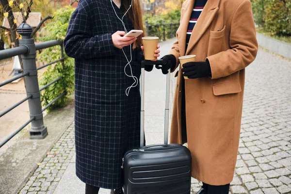 Feche duas garotas elegantes com café para ir e mala no parque da cidade — Fotografia de Stock