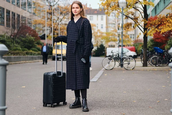 Стильная девушка с чемоданом ждет такси у дороги на уютной городской улице — стоковое фото