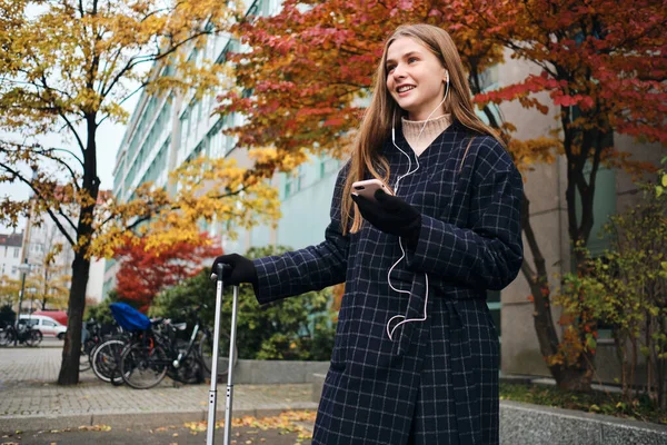 Menina muito alegre em fones de ouvido alegremente ouvir música no celular esperando na rua com mala — Fotografia de Stock