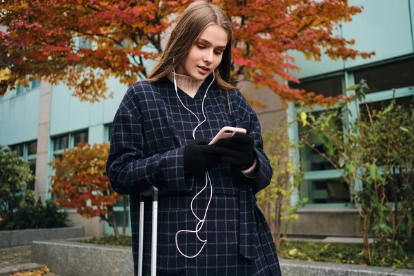 Atrakcyjne stylowe dziewczyny w słuchawkach za pomocą telefonu komórkowego stojącego na ulicy miasta z walizką — Zdjęcie stockowe