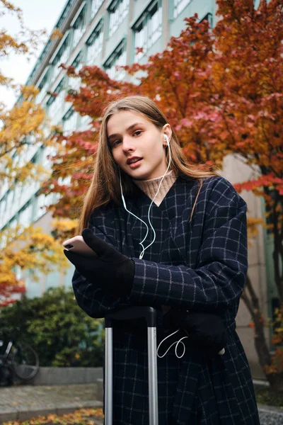 Όμορφη casual κορίτσι στα ακουστικά με κινητό τηλέφωνο με αυτοπεποίθηση κοιτάζοντας στην κάμερα στο δρόμο με τη βαλίτσα — Φωτογραφία Αρχείου
