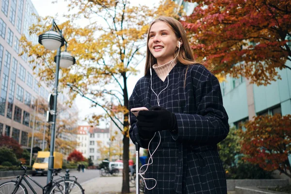 Krásná stylová dívka ve sluchátkách s mobilním telefonem šťastně čeká taxi na útulné městské ulici — Stock fotografie