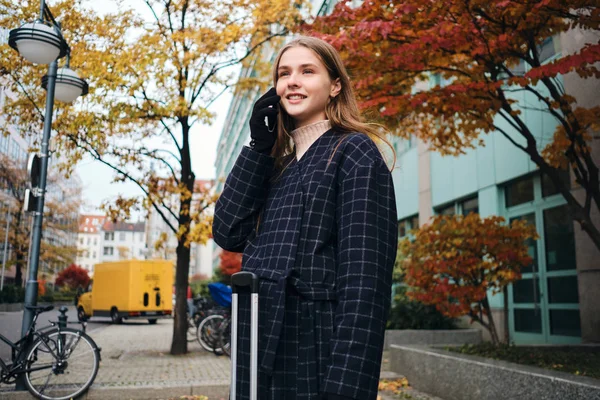 Красивая стильная девушка счастливо разговаривает по мобильному телефону стоя на городской улице с чемоданом — стоковое фото