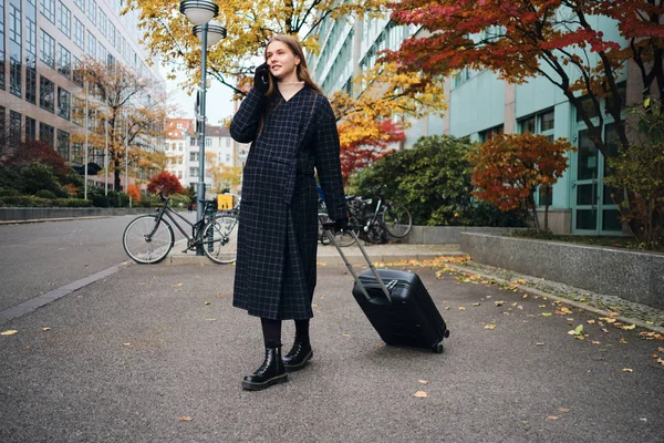 Menina casual atraente feliz falando no celular esperando táxi com mala na rua da cidade — Fotografia de Stock